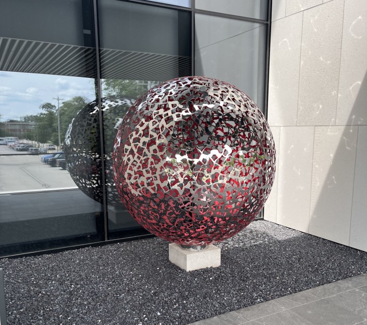 A stainless steel Geminus contemporary sculpture with red accents stands on a small plinth by a modern building.