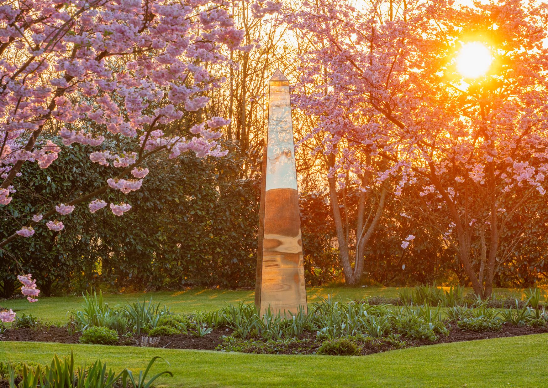 A mirrored obelisk stands among blossoming cherry trees at sunset in the tranquil garden sundial
