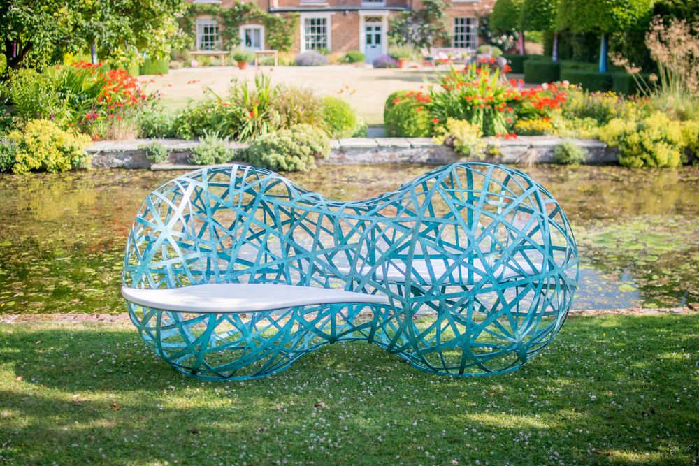 Modern, blue woven outdoor bench on grass—perfect for garden sculpture ideas—in a lush garden with pond and house.