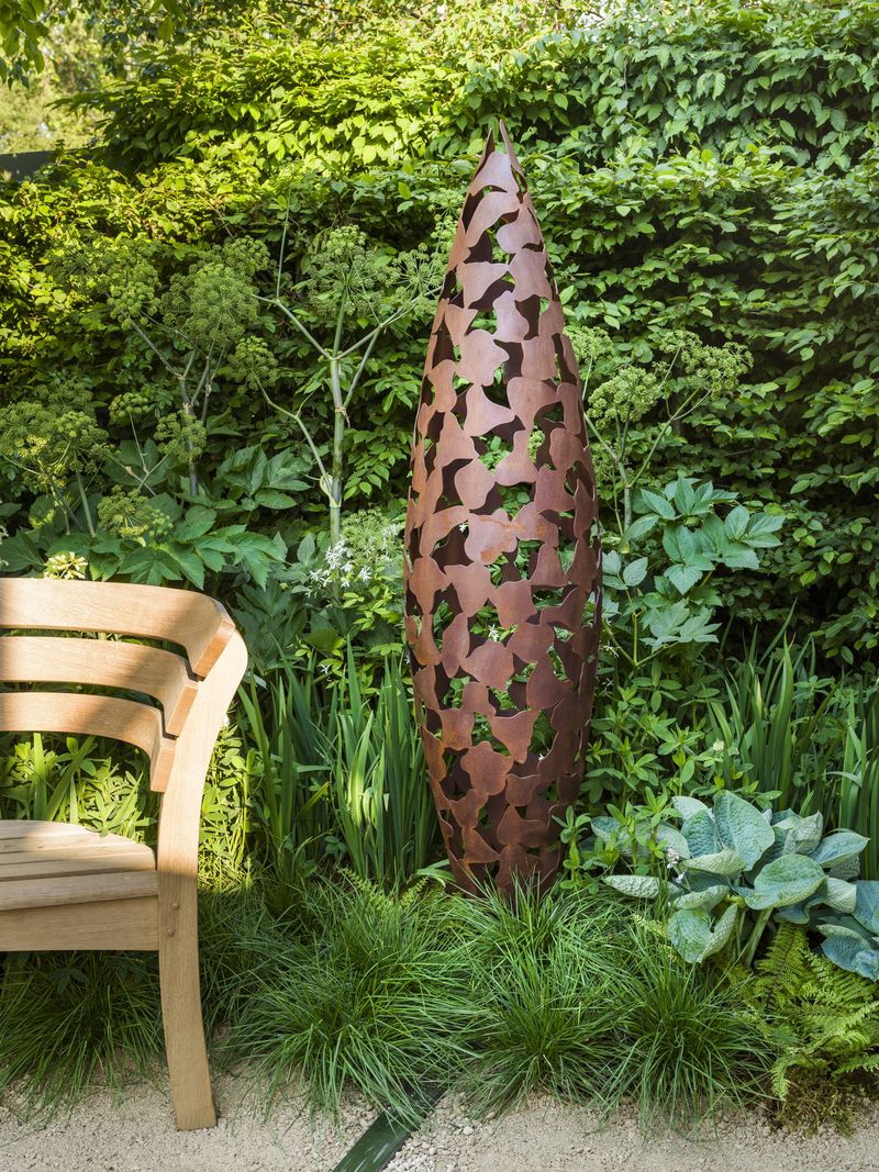 Tall, brown metal sculpture with leaf cut-outs stands in a lush courtyard garden, inspiring new courtyard garden ideas.