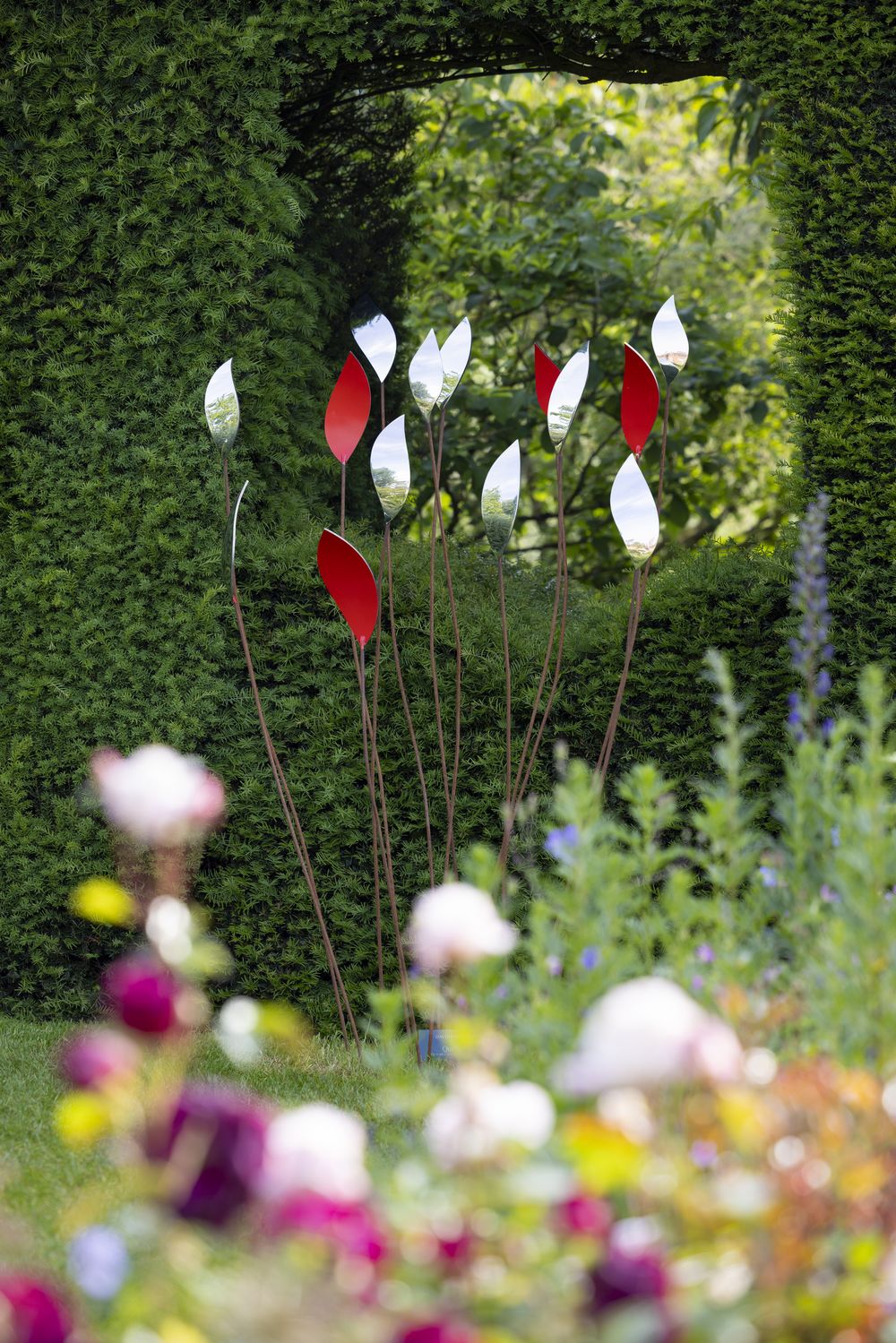 A group of tall, slender quiver organic sculptures with silver and red leaf-shaped tops stands in front of a large green hedge with a rectangular opening, surrounded by colourful, blurred flowers and plants in this striking garden design.