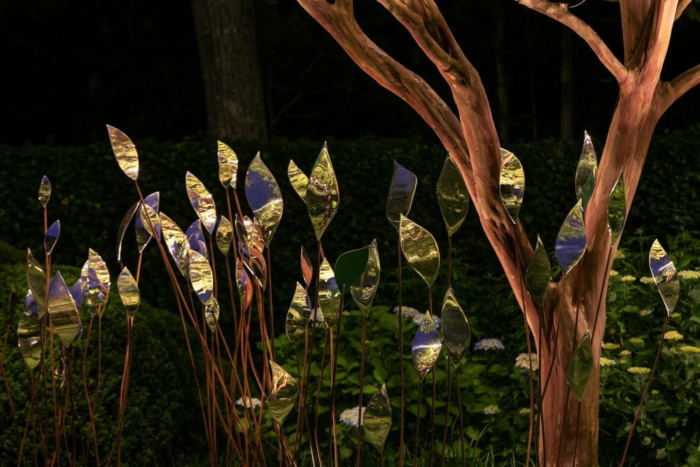 Quiver stainless steel leaf sculptures reflect light beside a bronze tree in a lush green garden.