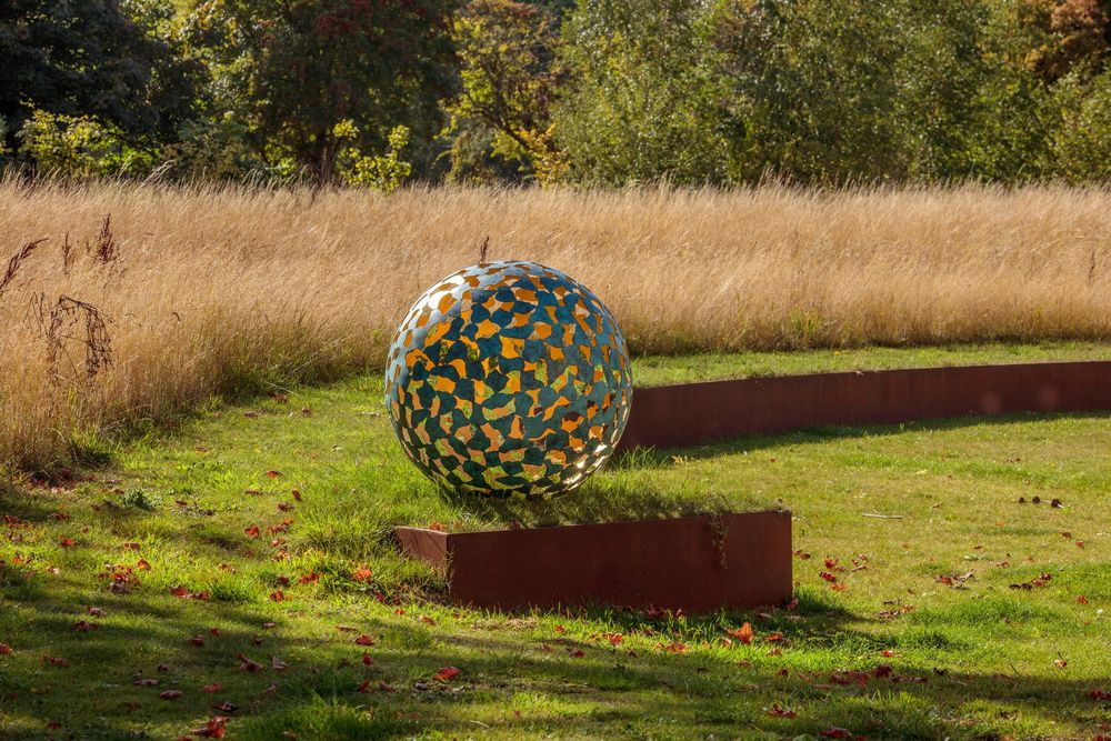 large bronze-colored sphere sculpture on a low, rusted corten steel retaining wall in a grassy field with tall yellow grass in the background