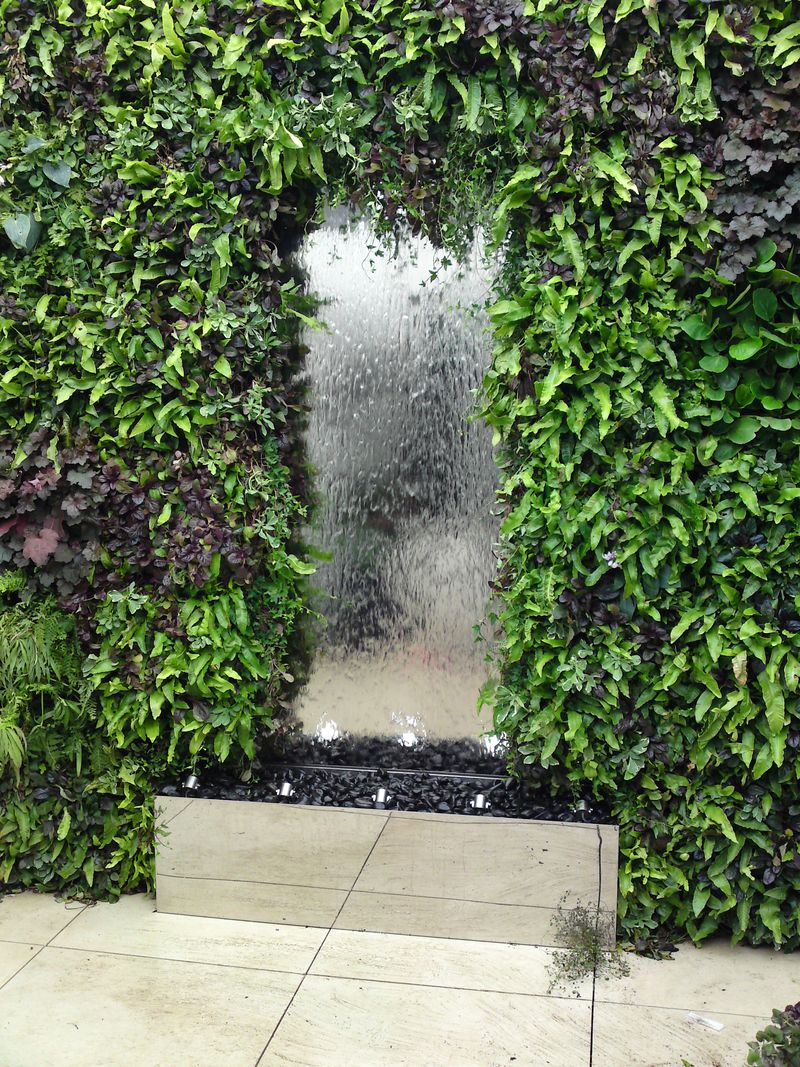 large stainless steel water feature with water cascading down a glass panel into a lush green living wall and stone basin.