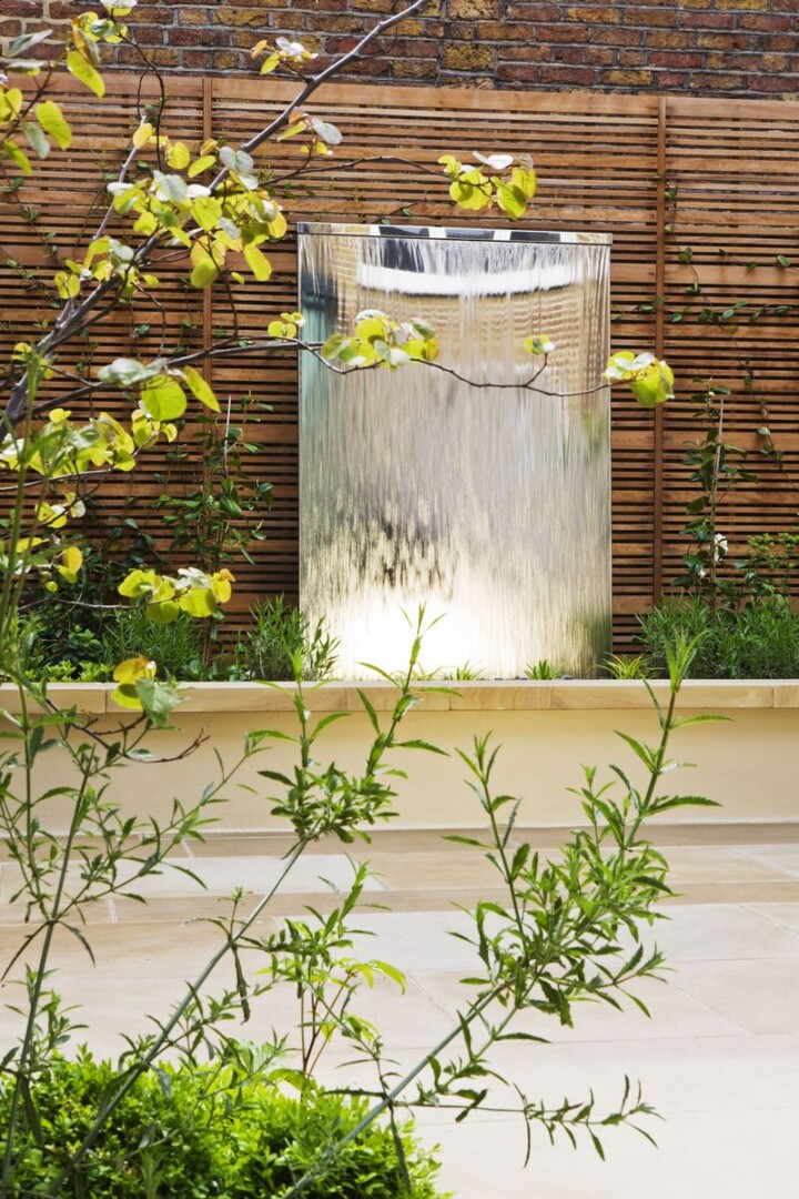 Modern garden with a large stainless steel water feature, wooden fencing, lush plants, and stone paving in the foreground.